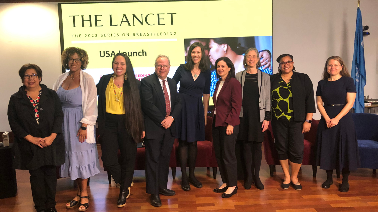 Nine individuals standing in a horizontal line smiling for a group photo. Each individual is wearing professional attire. They pose in front of a screen that reads “THE LANCET The 2023 Series on Breastfeeding USA Launch”