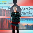 A person stands in front of a projected image featuring text, graphics, and the UC Davis Global Migration Center logo. The background shows silhouettes of people near a border fence, accompanied by Spanish text, including "Humanizando la Deportación" and "Visualizando el Asilo." The projection overlaps onto the person’s clothing and body, integrating them into the imagery.