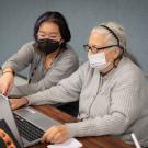 Student sitting side by side with an older adult. The student points to something on the laptop screen that they are both looking at.