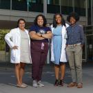 A group of four women stand together in front of a medical office building. Two wear lab coats and one wears scrubs. One has a stethoscope. All are smiling and looking directly at us.
